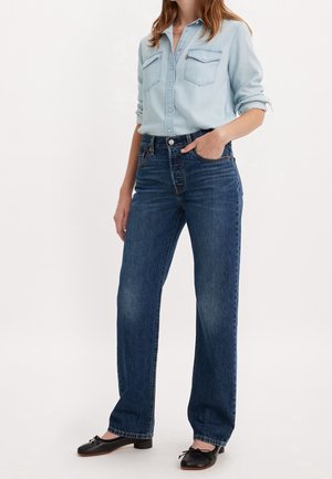 Kvinde iført lyseblå skjorte med knapper og opkrogede ærmer, mørkeblå jeans med lige ben og sorte balletsko, hånd i lommen, stående.