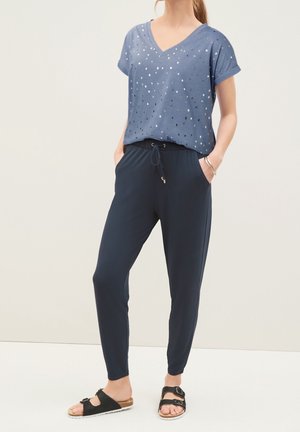 Mujer con blusa azul de manga corta con estampado de estrellas y cuello en V, pantalones azul marino con cordón y sandalias negras con hebilla, de pie con las manos en los bolsillos.