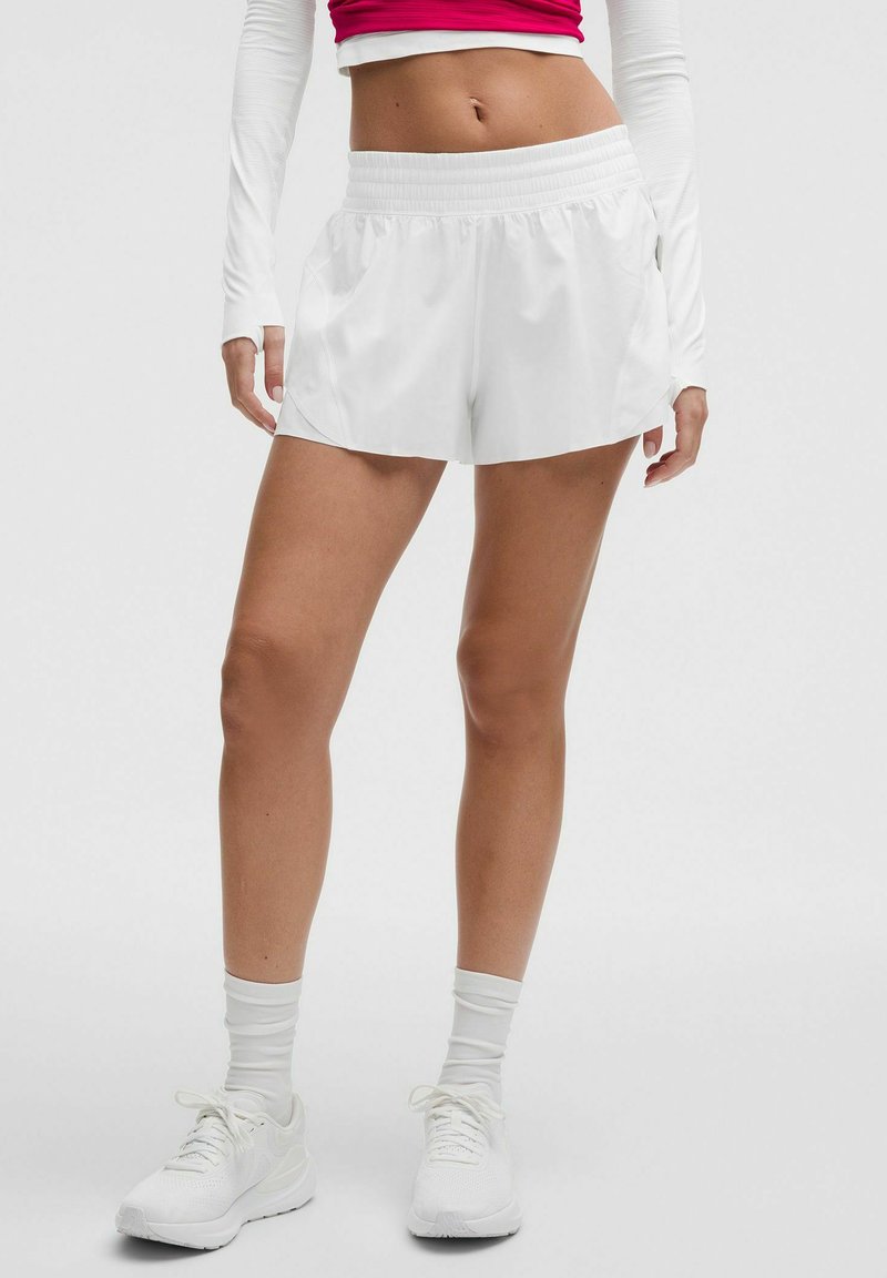 Femeie purtând pantaloni scurți atletici albi, adidași albi, șosete albe și un tricou roșu crop cu mâneci lungi, stând pe un fundal simplu.