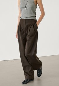 Ruskeat leveät housut, joissa on pleatit, valmistettu sileästä kankaasta. Mallilla on päällään harmaa toppi ja mustat loaferit. Minimalistinen muotoilu.