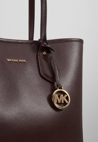 Sac cabas Michael Kors en cuir marron foncé avec deux poignées et un pendentif circulaire doré avec le logo MK suspendu à une des poignées.