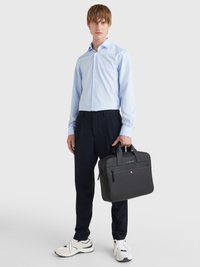 Malette en cuir noir avec deux poignées, texture lisse, poche zippée à l'avant et détail de logo subtil. Le modèle porte une chemise bleu clair et un pantalon sombre.