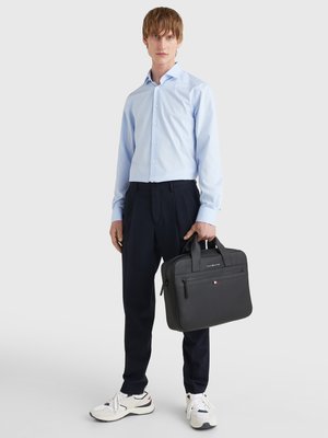 Black leather briefcase with two handles, smooth texture, front zippered pocket, and subtle logo detail. Model wears a light blue shirt and dark pants.