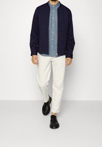 Giubbotto a scacchi blu navy sopra una camicia azzurro chiaro con bottoni, abbinato a pantaloni bianchi e scarpe nere con suole tonde e lacci.
