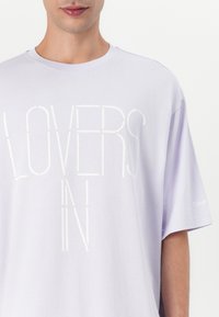 Lila bavlněná tričko s velkým bílým текстem "LOVERS IN," krátkými rukávy a malým logem na levém rukávu.
