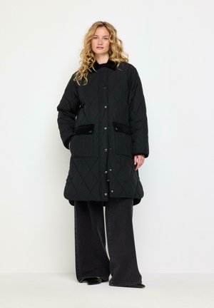 Vrouw met krullend blond haar die een lange zwarte gewatteerde jas met zakken en wijde zwarte broek draagt tegen een effen witte achtergrond.