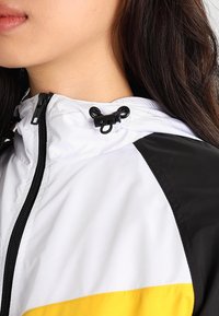 Bílá a černá kapucová bunda se žlutým akcentem; má nastavitelné stahování v oblasti krku, hladkou texturu a zapínání na zip.