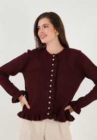 Burgundijas krāsas kardigāns, izgatavots no mīksta materiāla, ar volānveida apakšmalu, pogu aizdari un garām piedurknēm ar volānveida aprocēm.