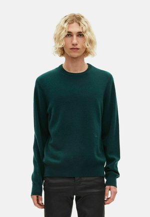 Jongere met krullend blond haar, gekleed in een donkergroene trui met lange mouwen en zwarte broeken, staand tegen een effen lichte achtergrond.