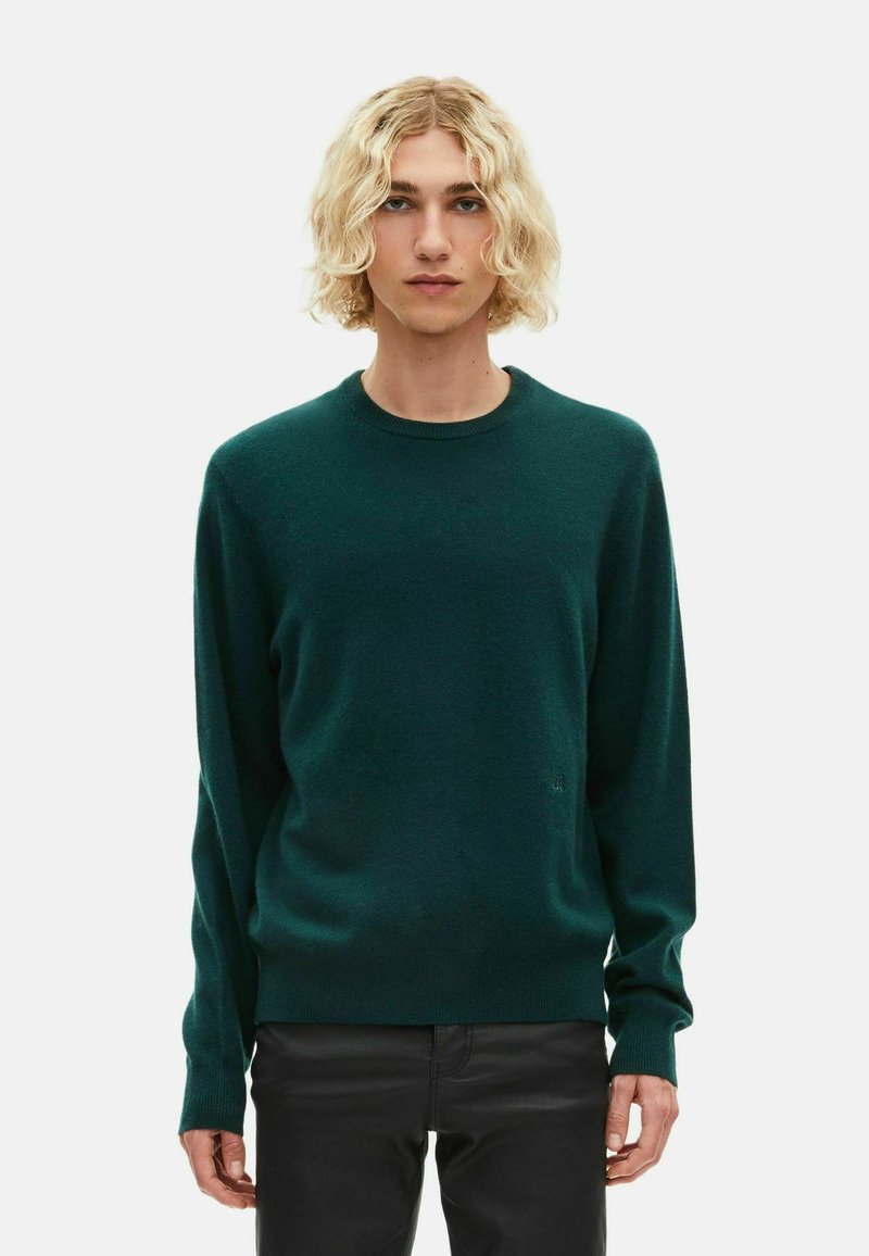 Młoda osoba z kręconymi blond włosami, ubrana w ciemnozielony sweter z długim rękawem i czarne spodnie, stojąca na tle jasnego, jednolitego tła.