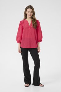 Blouse rose avec un décolleté en V et des motifs texturés, dotée de manches bouffantes. Associée à un pantalon noir évasé et des sandales.