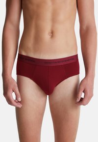 Torso maschile che indossa slip color bordeaux con logo "United Colors of Benetton" sulla cintura su sfondo bianco.