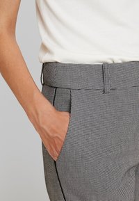 Spodnie w wysokim stanie w czarno-białą kratę z boczną kieszenią i eleganckim czarnym wykończeniem. Tkanina wydaje się gładka i dobrze ustrukturyzowana.