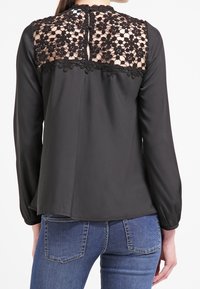 Blouse noire à manches longues, avec un dessus en dentelle au dos et une fermeture à bouton au cou. Tissu lisse avec une silhouette fluide.
