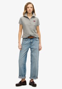 Grå polo-shirt med opadstående krave, der har et lille logo. Sammen med lyseblå, løstsiddende jeans og mørkebrune sko.