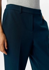 Gros plan sur une personne portant un pantalon tailleur bleu marine avec des passants de ceinture et un haut en maille gris clair rentré.