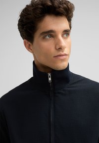 Schwarze Zip-Jacke aus strukturiertem Stoff, mit hohem Kragen und silbernem Reißverschluss. Das Material wirkt leicht und atmungsaktiv.