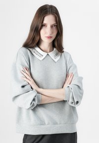 Grå sweatshirt laget av myk stoff med hvit krage og metalliske detaljer. Løs passform og mansjetterte ermer.