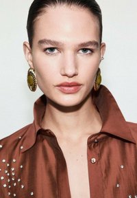 Camisa de satén marrón con cuello y adornos decorativos. La modelo lleva grandes pendientes dorados y verdes. Piel clara, maquillaje suave.