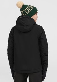 Zwarte geïsoleerde jas met een capuchon, met een gladde textuur. Een groene gebreide beanie met een pompon en logo-detail wordt op het hoofd gedragen.