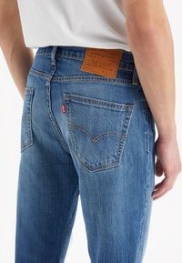 Blue denim jeans with a straight-leg design, featuring a leather brand tag, contrasting stitching, and back pockets with a signature logo detail.