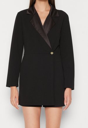 Frau trägt ein schwarzes Blazer-Kleid mit einem Satin-Revers und einem einzigen goldenen Knopf, steht vor einem einfarbigen Hintergrund.