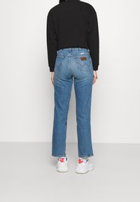 Jeans blu a gamba dritta con vita alta, con una toppa in pelle marrone sul retro, abbinati a una camicia nera e sneakers bianche.