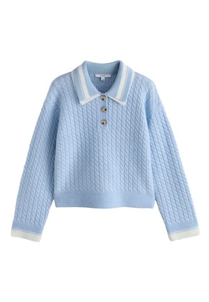 Mladá žena v svetlomodrom pulóvri, bielych širokých nohaviciach a bielych topánkach, usmievajúca sa a pózujúca s rukami zdvihnutými pred bielou stenou.