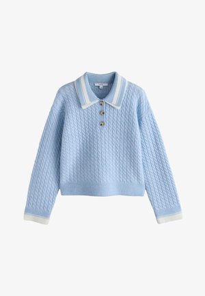 Giovane donna con maglione azzurro chiaro, pantaloni bianchi a gamba larga e scarpe bianche, sorridente e che posa con le braccia alzate contro una parete bianca.