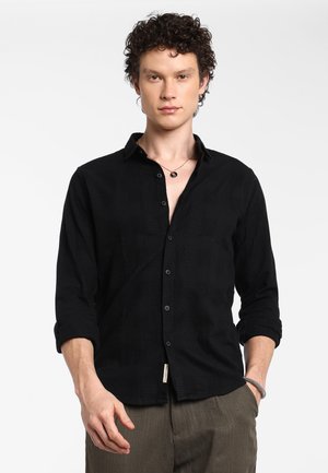Zwarte button-up blouse met een gestructureerd patroon, lange mouwen opgerold en een label op de zak. Draagt een relaxte fit broek.