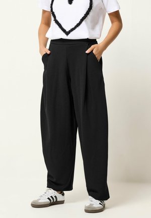 Personne portant un pantalon noir ample avec des poches latérales, des baskets blanches avec des rayures noires, et une chemise blanche avec un grand cœur noir texturé.
