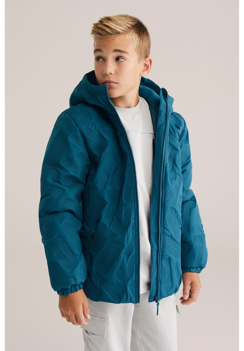 Chaqueta acolchada en color verde azulado con patrones geométricos, cierre de cremallera y capucha ajustable adjunta. Textura suave con puños elásticos.
