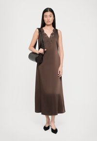 Rochie fără mâneci, de culoare maro, cu detalii din dantelă la decolteu, fabricată dintr-un material neted. Modelul ține în mână o geantă rotundă neagră și poartă pantofi negri.