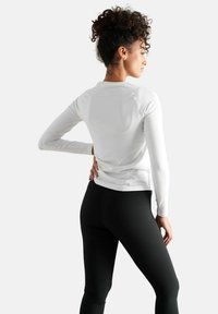 Hvid langærmet top med rund halsudskæring, tætsiddende pasform og sømløs design, båret med sorte leggings. Glat tekstur, minimale detaljer.