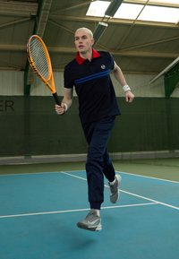 Kék és fekete sportöltözet, piros gallérral, amely egy pályán fut, narancssárga teniszütőt tart a kezében, szürke sportcipőben.