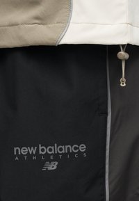 Κοντινό πλάνο μαύρου και μπεζ αθλητικού μπουφάν με το λογότυπο New Balance και ρυθμιζόμενο κορδόνι στο επάνω δεξιό μέρος.