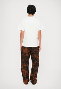 Weißes kurzärmliges T-Shirt kombiniert mit braunen, graphisch gemusterten weit geschnittenen Hosen, die eine Mischung aus dunklen Schattierungen und Texturen aufweisen.