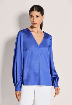 Felle blauwe satijnen blouse met een V-hals, lange pofmouwen en manchetten met knopen; gladde textuur en losse pasvorm, gecombineerd met witte broek.
