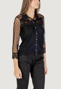 Blusa nera con maniche trasparenti a pois, dettagli in pizzo e chiusura frontale con bottoni. Presenta un orlo ondulato e motivi floreali intricati.