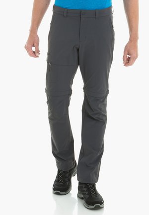 Mann trägt dunkelgraue Zip-Off-Wanderhose und schwarze Wanderschuhe, steht mit entspannt an den Seiten hängenden Händen.