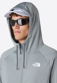 Lys grå zip-up hoodie med en struktureret overflade, justerbare snore og hvidt logo på venstre bryst, båret med solbriller og en kasket.