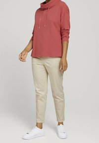 Czerwony, fakturowany top z kołnierzem typu komin i ściągaczami, zestawiony z beżowymi, zwężanymi spodniami i białymi trampkami, noszony przez osobę.