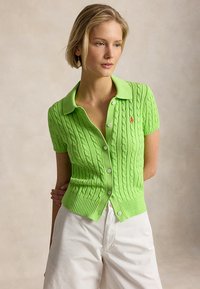 Nainen, joka pitää yllä kirkkaanvihreää lyhythihaista palmikkoneulottua neuletakkia, jossa on valkoiset napit, sekä valkoisia housuja, poseeraa neutraalia taustaa vasten.