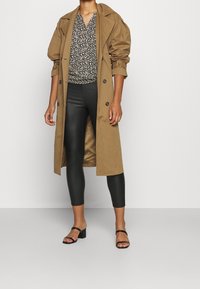 Beige Trenchcoat mit großen Knöpfen, schwarze Bluse mit floralem Muster, schwarze Leggings und schwarze Sandalen mit Absatz und Riemchen. Glatte Texturen und geschichtetes Design.