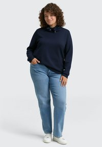 Marineblauwe pullover met een hoge kraag en trekkoorddetails, gecombineerd met lichtblauwe rechte jeans en witte sneakers. Zachte stoftextuur.