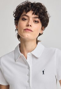 Polo shirt blanc en tissu texturé, avec un col classique et un rabat de boutons. Sur la poche gauche, se trouve un petit logo d'un golfeur.