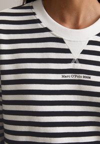 Nahaufnahme eines schwarz-weiß gestreiften Shirts mit einem weißen gerippten Kragen und "Marc O'Polo DENIM" auf der Brust gestickt.