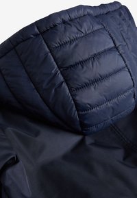 Marineblå dunjakke med hætte, som har quiltede mønstre, glat tekstur og mat finish, der går over i et lysere stof forneden.