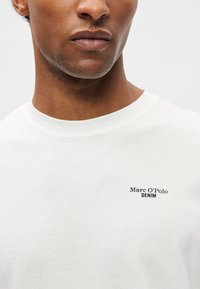 Balta kokvilnas T-krekls ar apaļo dekoltē, kuram ir neliels melns logo, kas lasāms "Marc O'Polo" un "DENIM" kreisajā krūtīs.