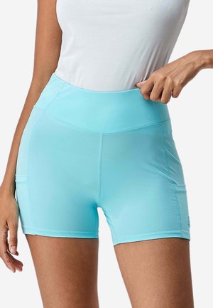 Donna che indossa pantaloncini sportivi a vita alta azzurri con tasche laterali e una canotta bianca senza maniche, che regola la cintura.